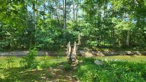 A forested area with a narrow path leading to a bridge over a calm body of water. - Hotel Pasteral (La Cellera de Ter)