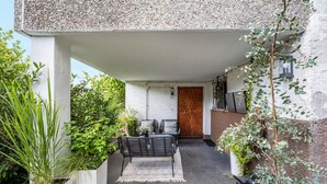 Un espacio de descanso cubierto al aire libre con un banco y un tapete estampado, rodeado de follaje y una pared cubierta de piedras.