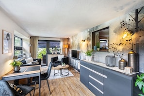 Un salon moderne avec un canapé, une table basse et un téléviseur. Il y a également un coin repas avec une table et des chaises. La pièce est dotée d’une grande fenêtre et offre une vue sur la verdure environnante.