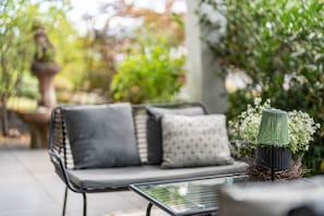 Un espace patio aménagé avec un canapé en métal noir, une table en verre et un pot de fleurs orné de fleurs blanches.