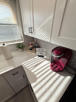 Una cocina con armarios blancos, una cafetera, un letrero que dice 'COFFEE TIME' y un ventanal con persianas.
