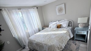 Um quarto com cama, mesa de cabeceira, luminária, janela com cortinas e uma obra de arte na parede.