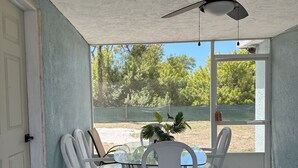 Quarto com ventoinha no teto, mesa de vidro, cadeiras brancas e vista para árvores e uma cerca lá fora.