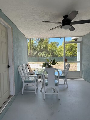 Habitación con ventilador de techo, una mesa de vidrio, sillas blancas y vista a árboles y una cerca afuera.
