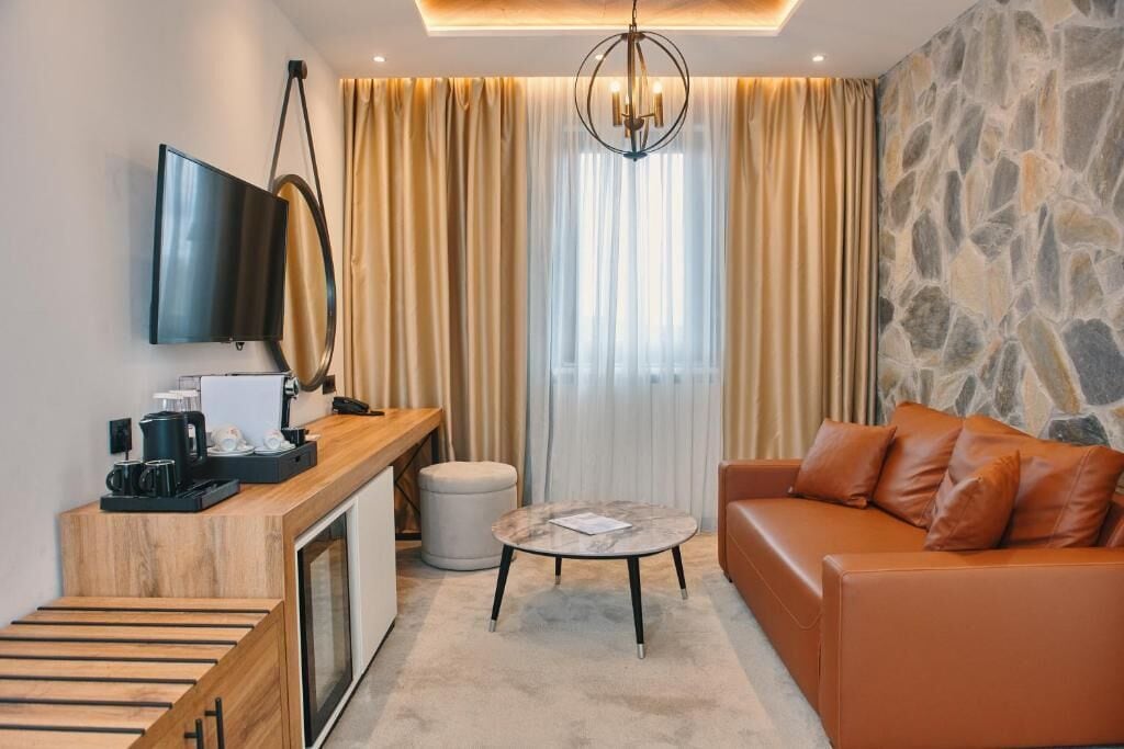 A modern hotel room with a wooden desk, a television, a stone wall, a leather sofa, and a round table.