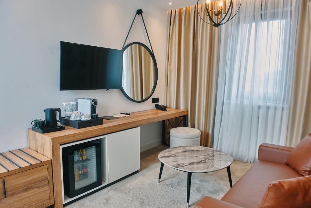 A modern hotel room with a kitchenette, a flat-screen TV, a round mirror, a sofa, and a coffee table.
