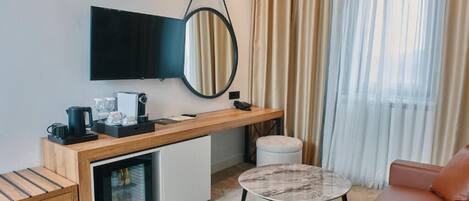 A modern hotel room with a kitchenette, a flat-screen TV, a round mirror, a sofa, and a coffee table.