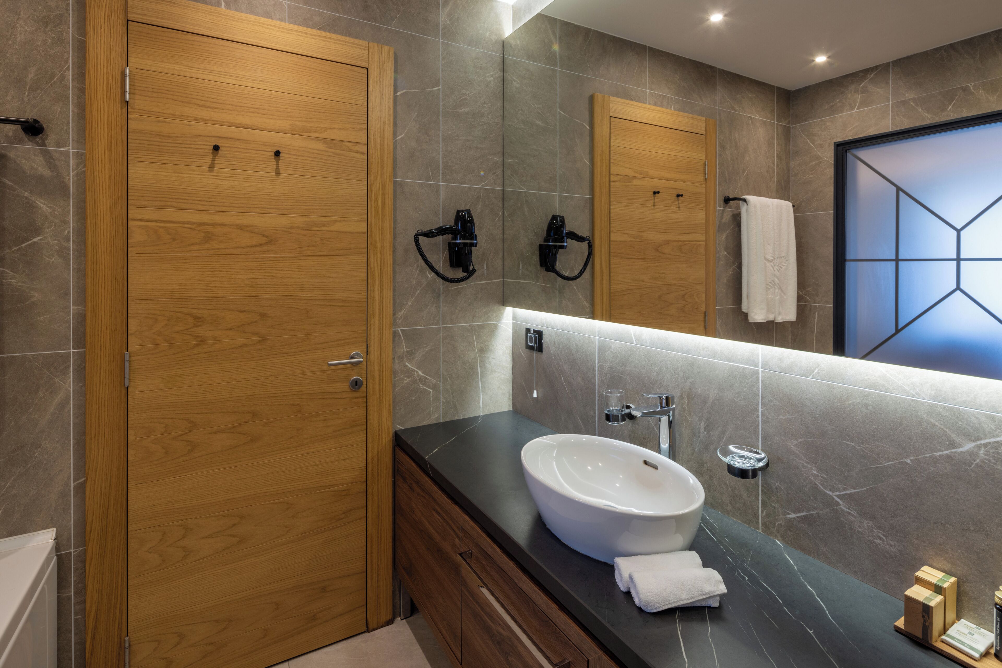 Une salle de bain moderne avec une porte en bois, un plan de travail noir, un lavabo blanc et un grand miroir.