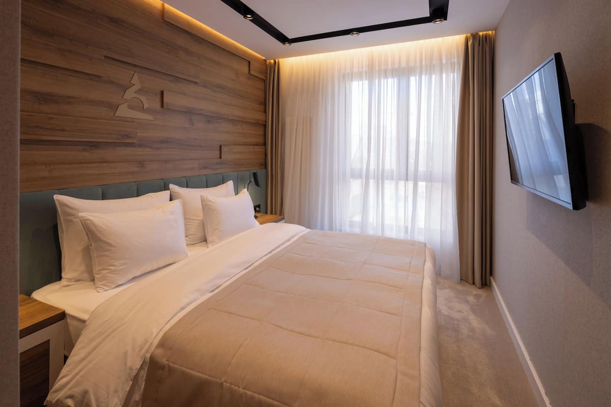 Une chambre d’hôtel moderne dotée d’un grand lit, d’une tête de lit en bois, d’un téléviseur à écran plat et d’une grande fenêtre avec des rideaux.