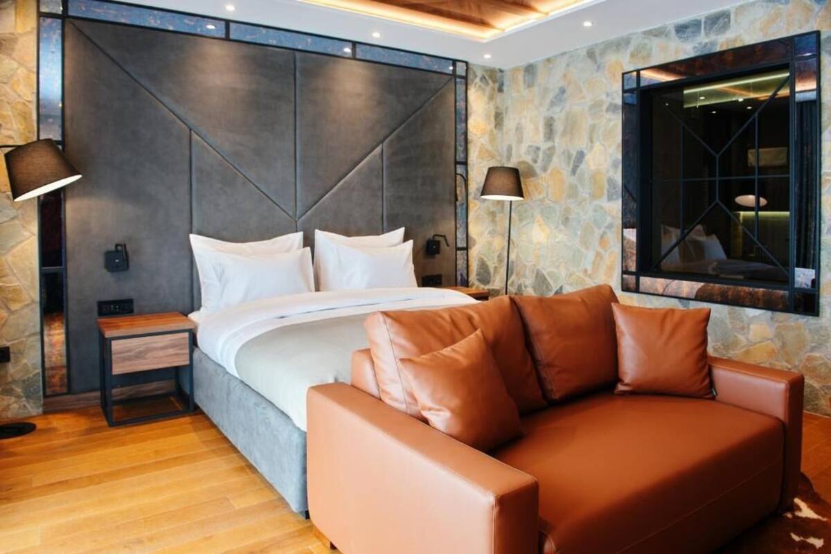 Une chambre à coucher moderne avec un grand lit, un canapé en cuir, des murs en pierre et une fenêtre dotée d’un cadre géométrique.