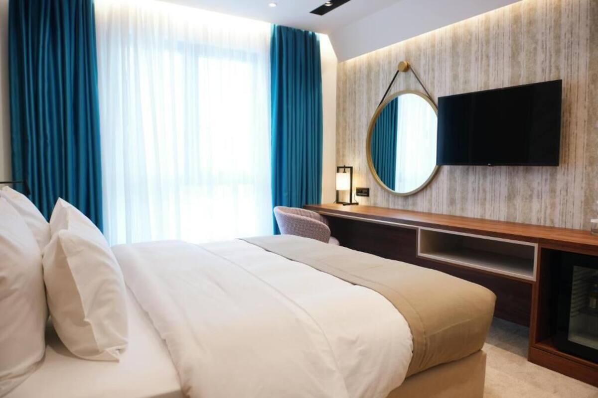 Une chambre d’hôtel avec un lit, un bureau, une télévision, un miroir et une fenêtre avec des rideaux.