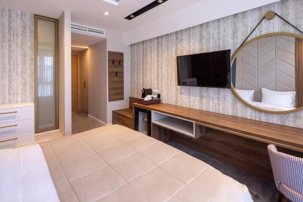 A modern hotel room with a large bed, a wooden console table, a round mirror, a flat-screen TV, and a built-in wardrobe.