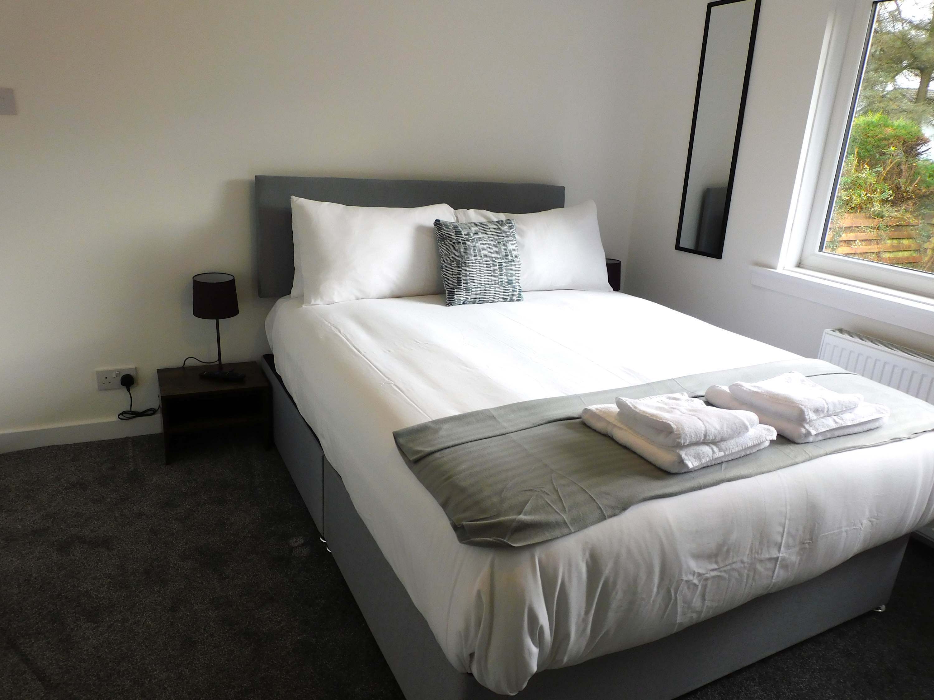A neatly made bed with white linens, a grey throw blanket, and folded towels. There is a bedside table with a lamp, a power outlet, and a window with a view of greenery outside.