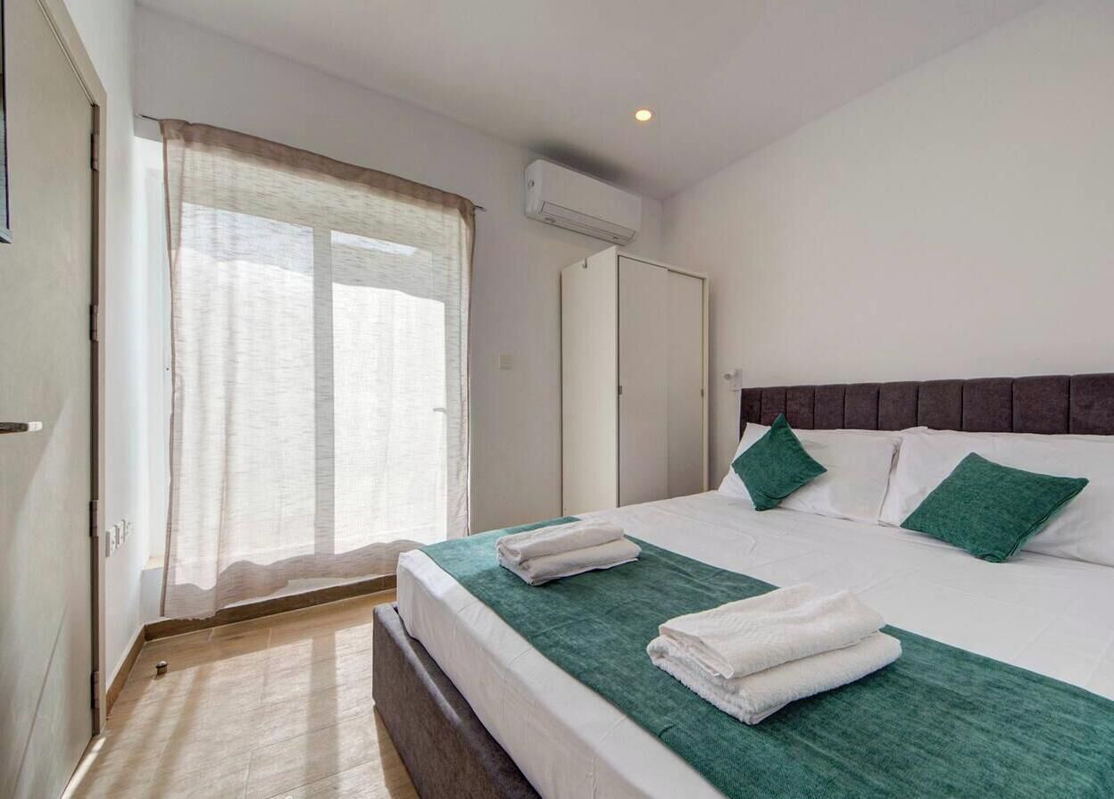 Une chambre à coucher avec un lit, des oreillers verts, une fenêtre avec des rideaux, un appareil de climatisation et une porte.