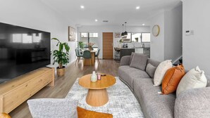 A modern living room with a flat-screen TV, a wooden coffee table, a grey sofa with decorative pillows, and a dining area in the background.