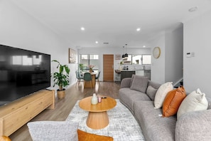Ein modernes Wohnzimmer mit Flachbildfernseher, einem Holztisch, einem grauen Sofa mit dekorativen Kissen und einem Essbereich im Hintergrund.