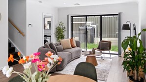 A modern living room with a grey sofa, a round coffee table, and a dining table. There is a staircase on the left, a sliding glass door leading to a balcony, and a potted plant in the foreground.