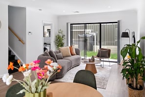Ein modernes Wohnzimmer mit einem grauen Sofa, einem runden Couchtisch und einem Esstisch. Links befindet sich eine Treppe, eine Schiebeglastür führt auf einen Balkon, und im Vordergrund steht ein Topfpflanzen.