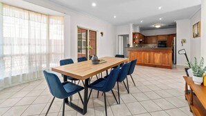 Un coin repas avec une table en bois et des chaises bleues, une cuisine avec des meubles en bois et une grande fenêtre ornée de rideaux légers.