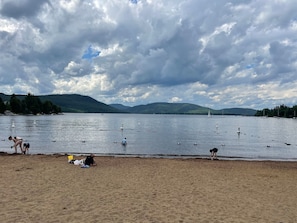 A beach with people swimming and sunbathing, a sandy shore, and a lake with boats.