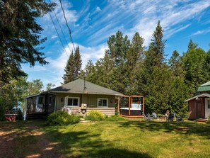 A single-story house with a porch, a hot tub, and a small shed in a wooded area. - Le Kaïa, Bord de L`eau, Plage, spa et foyer (Nominingue)