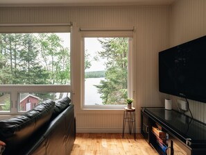 A living room with a black leather sofa, a television, and a window with a view of a lake and trees. - Le Kaïa, Bord de L`eau, Plage, spa et foyer (Nominingue)