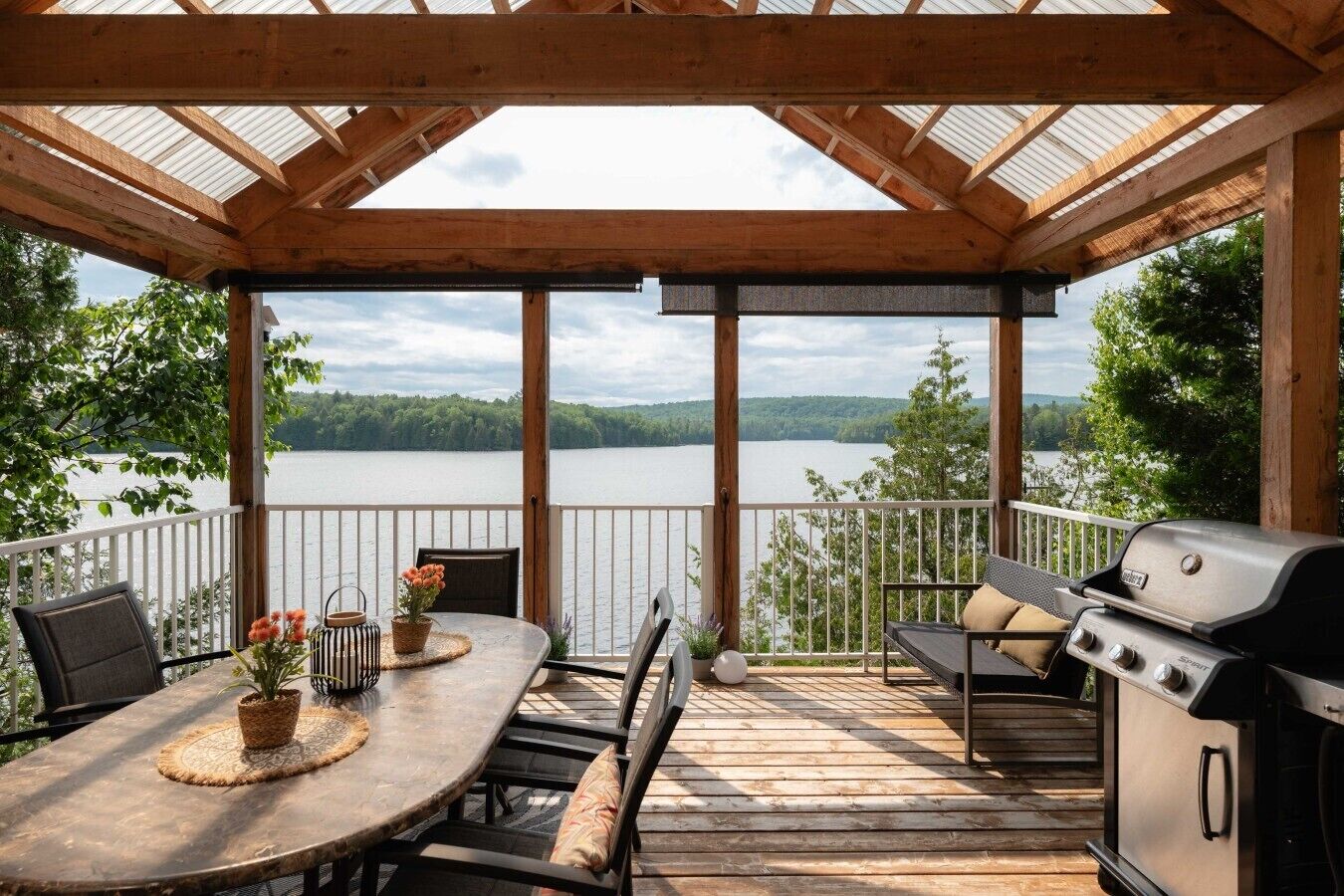 Une terrasse extérieure couverte, équipée d’une table et de chaises, d’un barbecue et offrant une vue sur un lac.