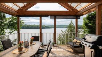 Une terrasse extérieure couverte, équipée d’une table et de chaises, d’un barbecue et offrant une vue sur un lac.