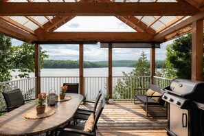 A covered outdoor deck with a table and chairs, a grill, and a view of a lake.