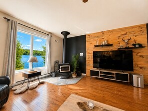 A living room with a large window, a fireplace, a TV, and a wooden wall. - La Baie la Vida,Bord de L`eau,Spa et foyer (Nominingue)