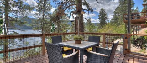 A deck with wicker chairs and a table, overlooking a lake with trees and a cabin in the background.