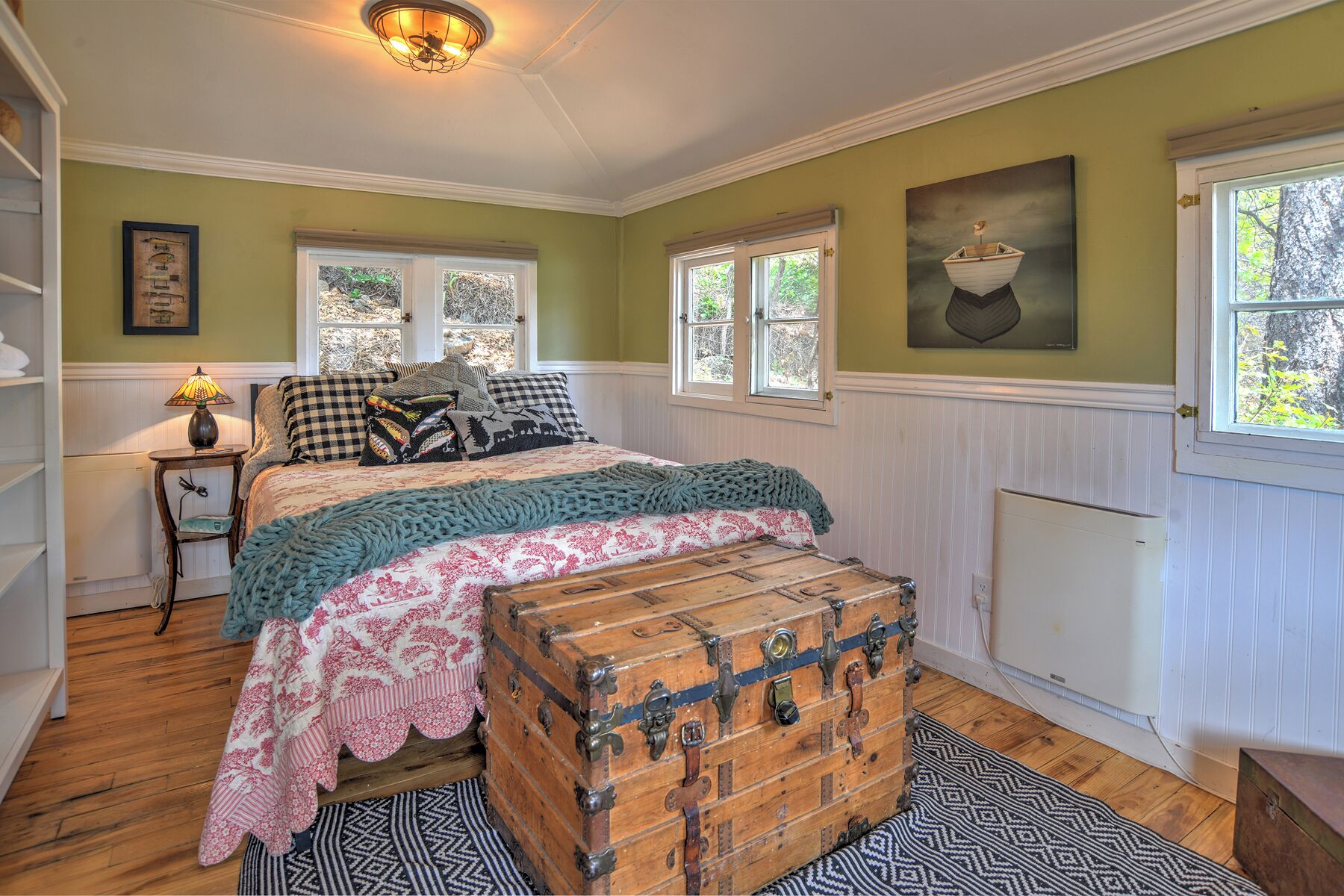 A bedroom with a bed, windows, a radiator, a chest of drawers, and a lamp.