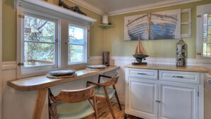 Une cuisine avec une table en bois, deux chaises, un meuble blanc et un tableau représentant un bateau accroché au mur.