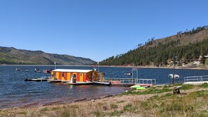 Un quai avec un petit bâtiment rouge et jaune, des bateaux et une colline boisée.