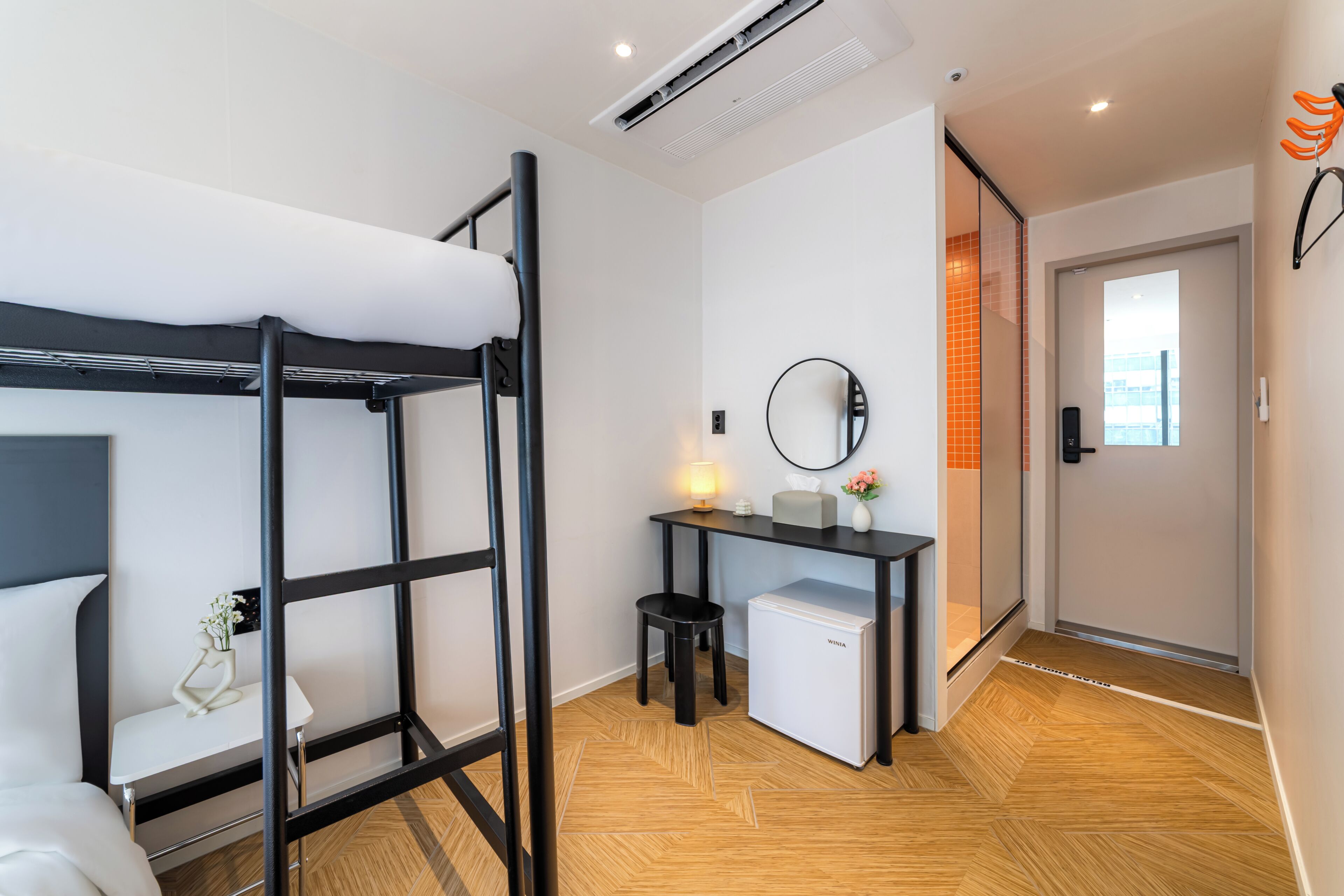 A modern room with a bunk bed, a small desk with a lamp, a refrigerator, and a mirror on the wall.