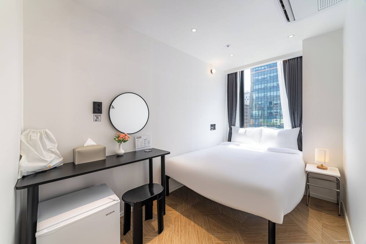 A hotel room with a bed, a desk with a computer, a small refrigerator, a mirror, and a window with curtains.