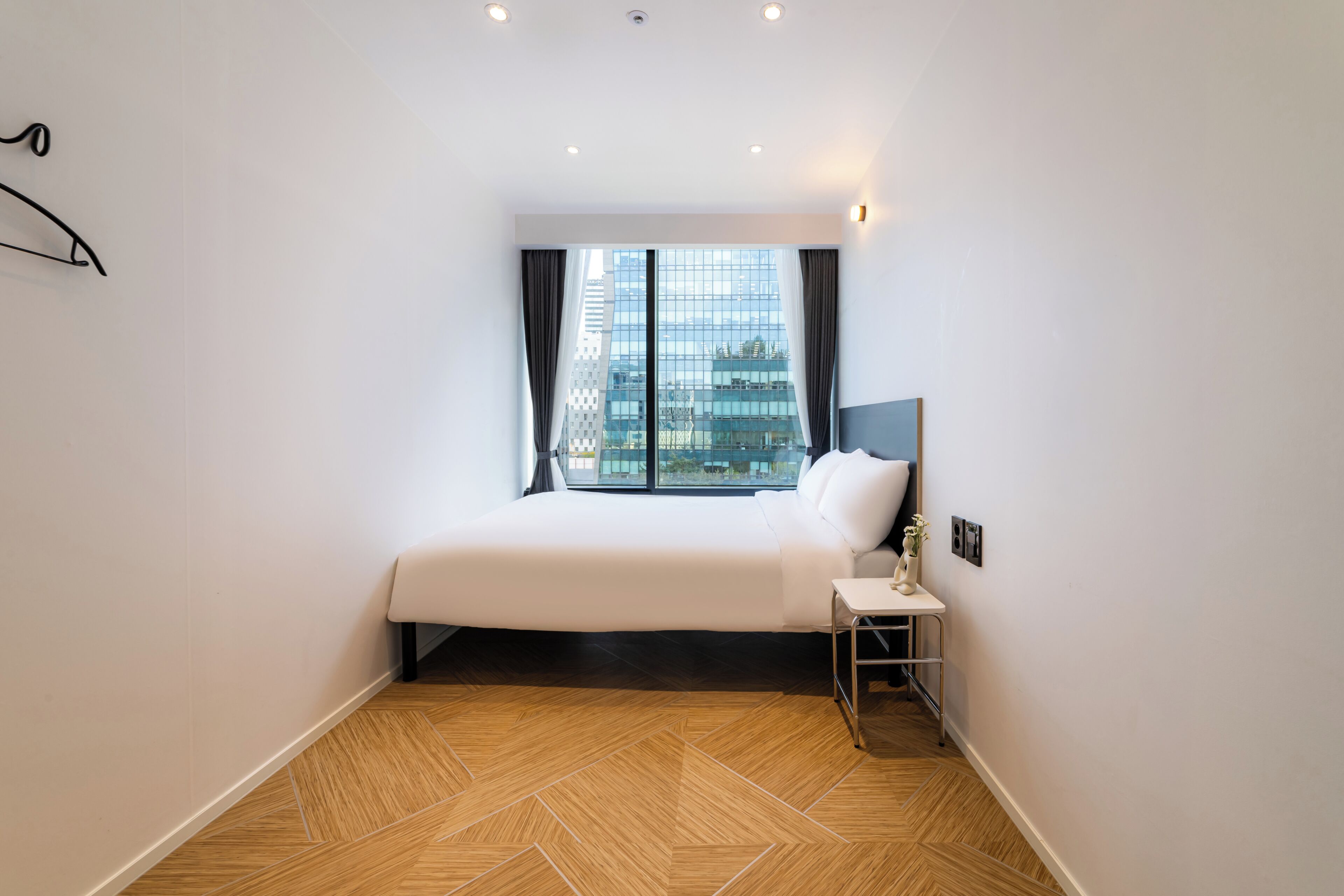 A hotel room with a large bed, a nightstand, a window with curtains, and a view of a cityscape.