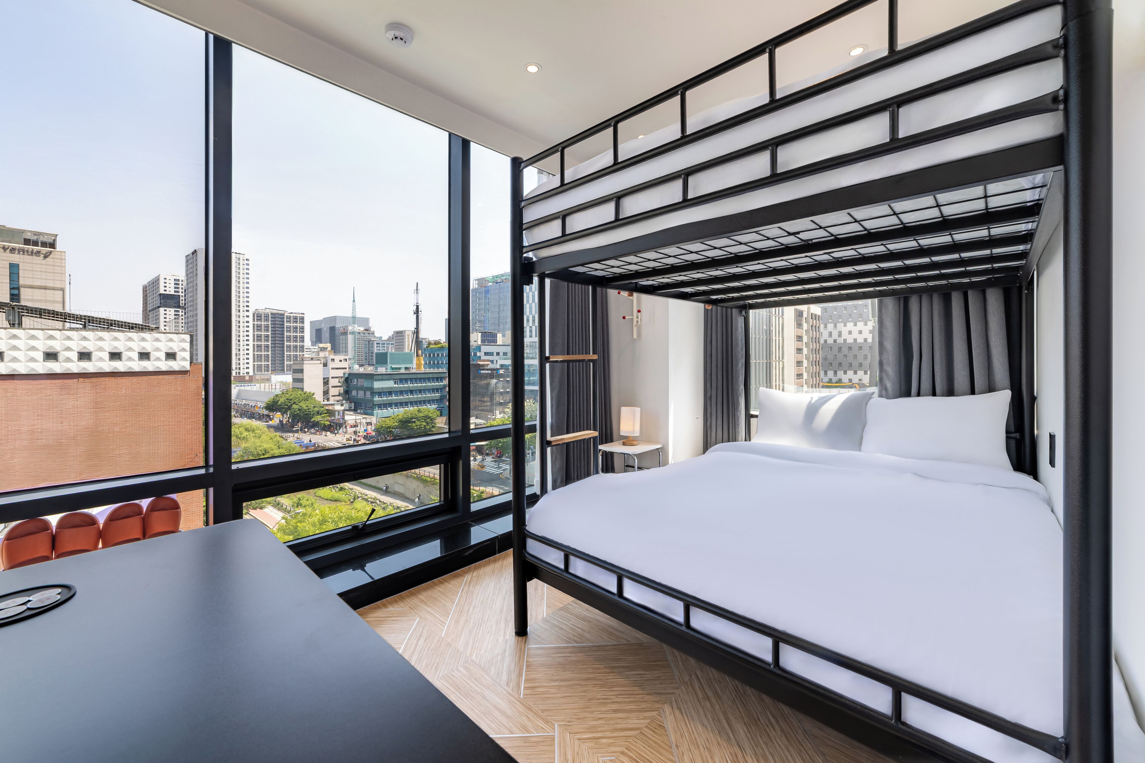 A modern hotel room with a bunk bed, large windows, a desk, and a view of the cityscape.