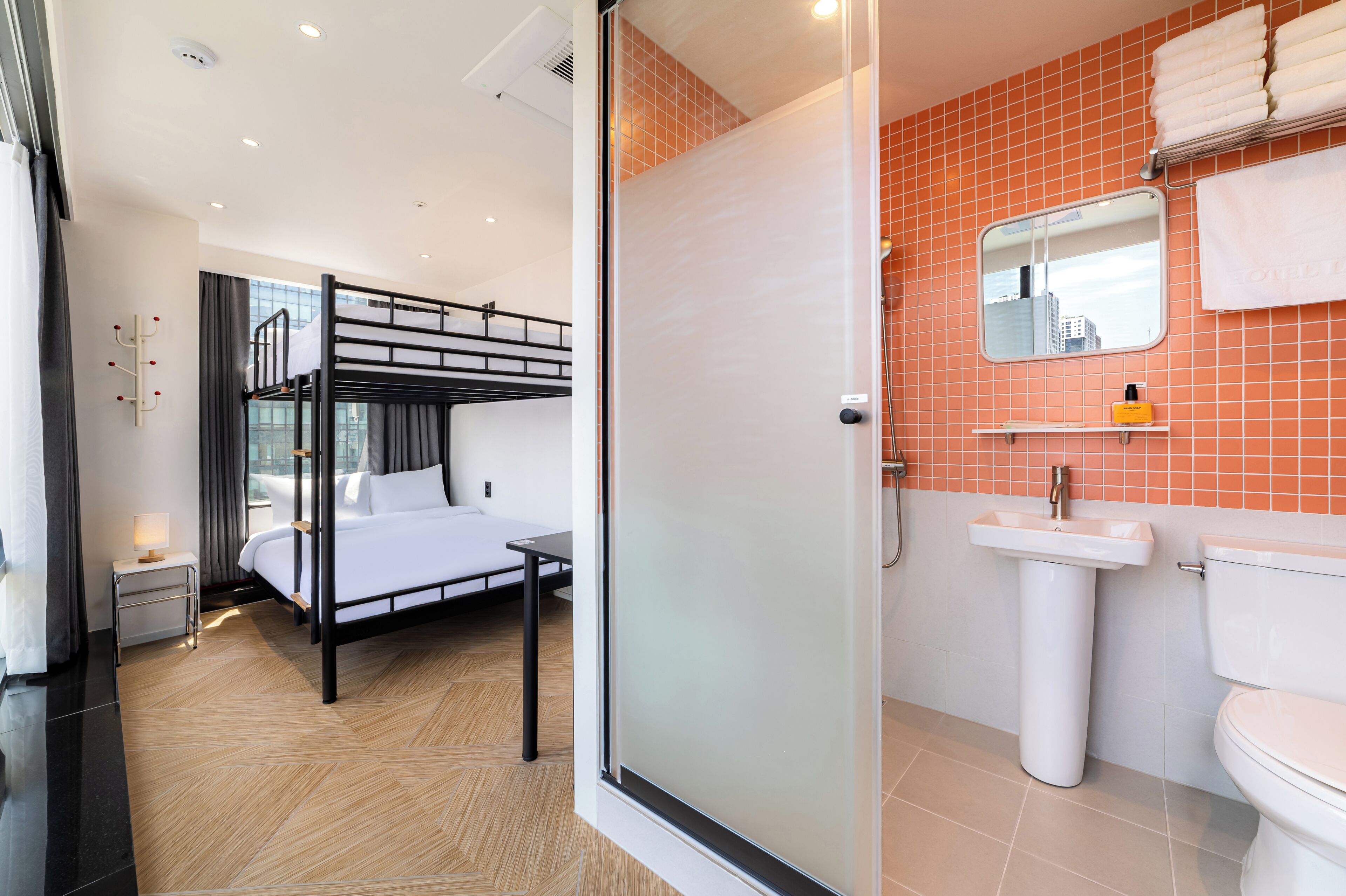 Una habitación de hotel moderna con una litera, una mesita de noche pequeña, cabina de ducha de vidrio, lavamanos blanco y un espejo.