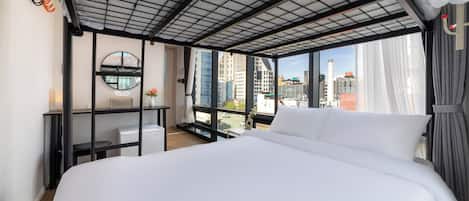 Una habitación de hotel moderna con una cama grande, un escritorio, una silla, un espejo redondo y vistas de la ciudad a través de amplias ventanas.