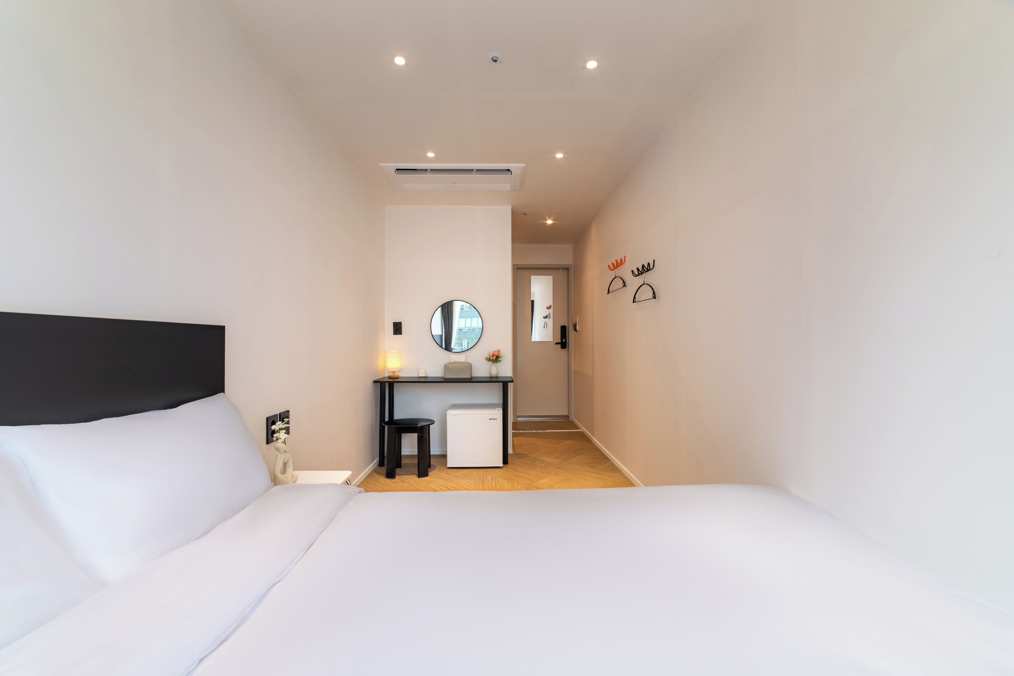 A bedroom with a bed, a desk with a chair, a small fridge, a mirror, and a door leading to another room.