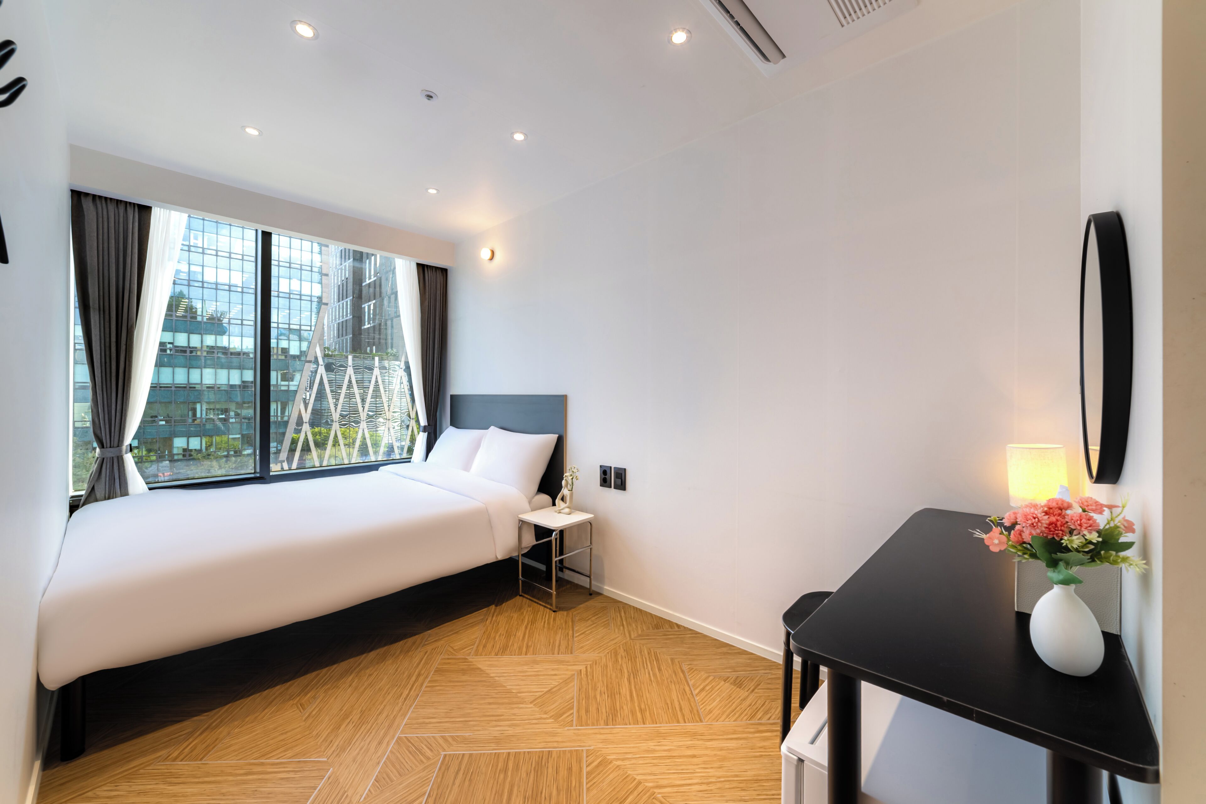 Habitación de hotel con una cama grande, una mesita de noche pequeña, un escritorio con un jarrón de flores y vistas a un edificio a través de la ventana.