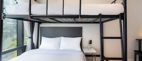 A bunk bed room with a view of a cityscape through large windows, a bedside table with a vase, and a small desk.