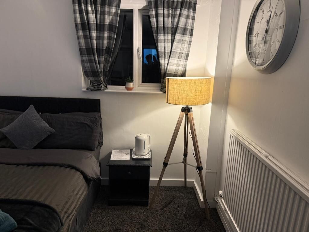 Ett sovrum med en säng, ett sängbord med en vattenkokare, en lampa på ett stativ, ett fönster med gardiner och en väggklocka.