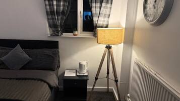 Un dormitorio con cama, una mesita de noche con hervidor, una lámpara sobre un trípode, una ventana con cortinas y un reloj de pared.