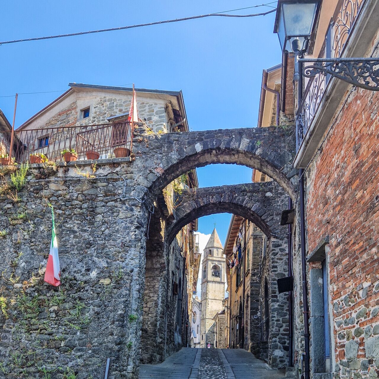 Una calle estrecha con edificios de piedra, una torre de iglesia a lo lejos y una bandera colgada a la izquierda.