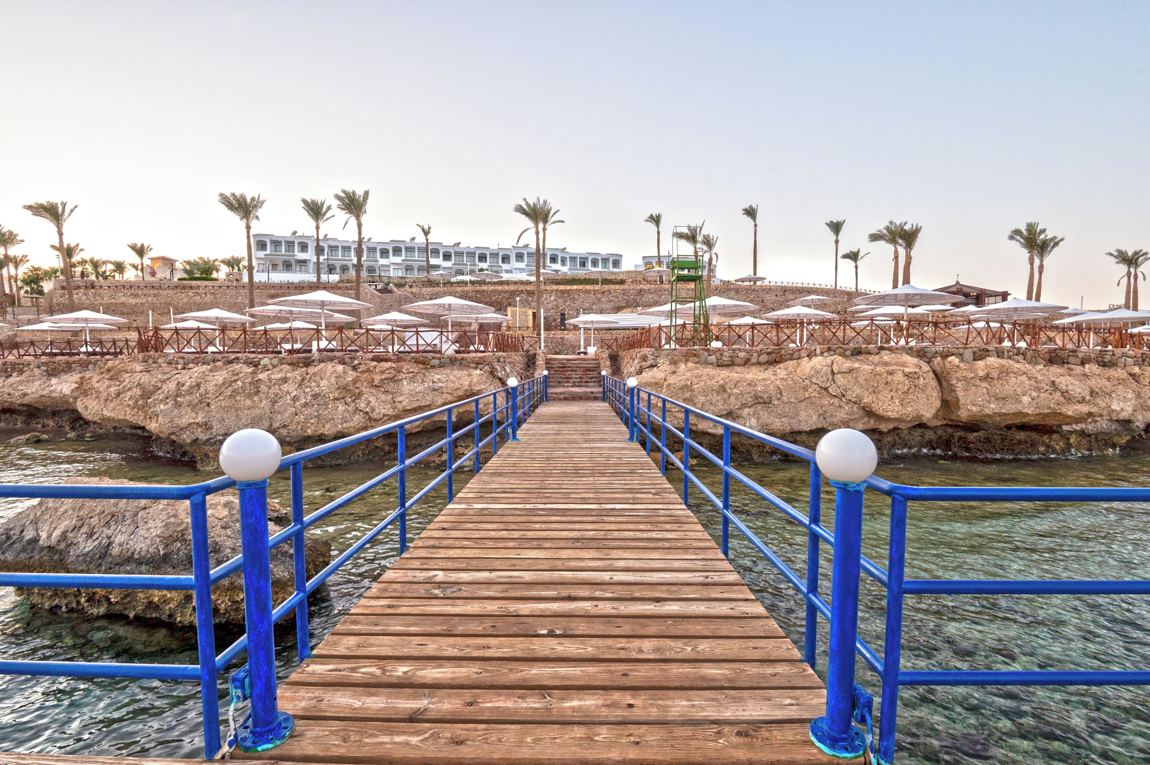 A wooden pier leading to a beach area with umbrellas and palm trees.