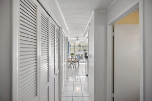 Een gang met witte wanden, een betegelde vloer en een deur die leidt naar een balkon met buitenmeubilair.