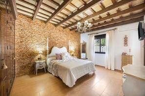 Een slaapkamer met een stenen muur, houten balken in het plafond, een bed met een witte kap, nachtkastjes met lampen en een dressoir.