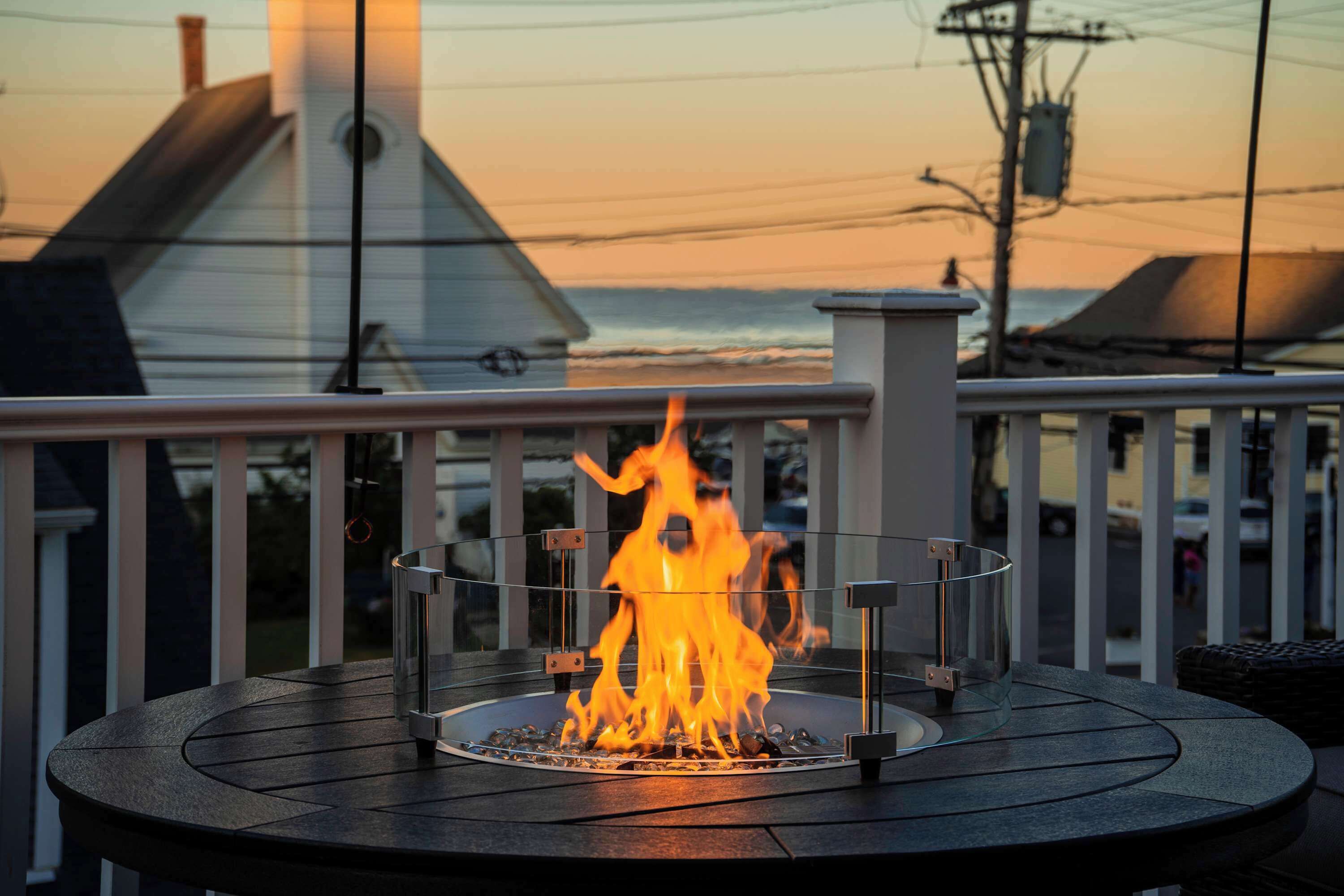 A circular outdoor seating area with a fire feature, surrounded by a railing with a view of the sky and buildings in the background.